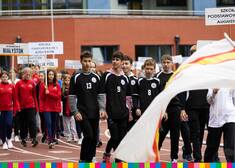 Młodzież w sportowych strojach przechodzi przez stadion trzymając tabliczki z nazwą szkoły
