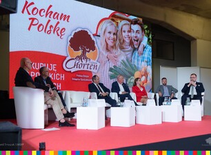 Sześciu mężczyzn i kobieta ubrana na czerwono siedzą na białych fotelach. W tle grafika Kocham Polskę