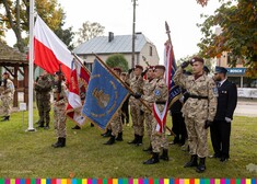 Młodzi ludzie w wojskowych strojach trzymają sztandar. W dalszym rzędzie mężczyźni z OSP