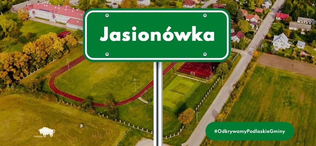 zielona tablica z nazwą miejscowości Jasionówka na tle zdjęcia miejscowości z lotu ptaka