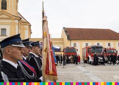 Po lewej stronie członkowie OSP trzymają sztandary, po prawej wozy strażackie i uczestnicy wydarzenia