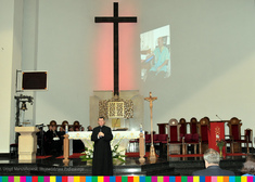 Kapłan stoi przed ołtarzem i przemawia. W tle widoczny krzyż oraz prezbiterium.