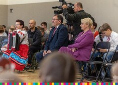 Wiesława Burnos wraz z dyrektorem szkoły Grzegorzem Nazarukiem oraz Panią trzymającą akordeon siedzą na widowni wraz z uczniami. W tle widać operatora z kamerą