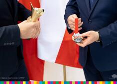 Dłonie trzymają medal i puchar na tle biało-czerwonej flagi