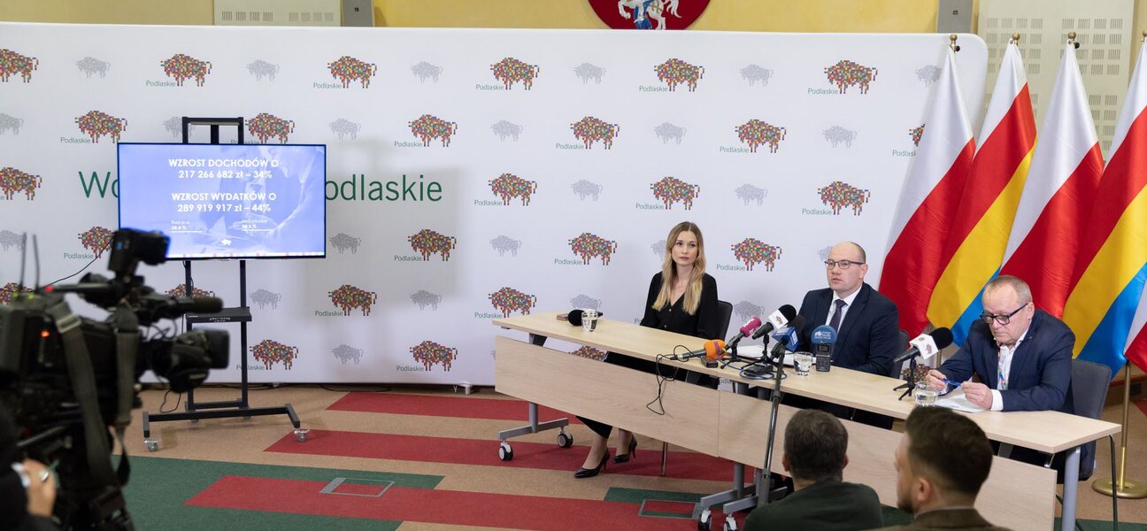 Marszałek, skarbnik i rzeczniczka prasowa siedzą na tle białej ścianki z logo województwa