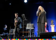 Dwie osoby na scenie, kobieta śpiewa a mężczyzna gra na saksofonie 