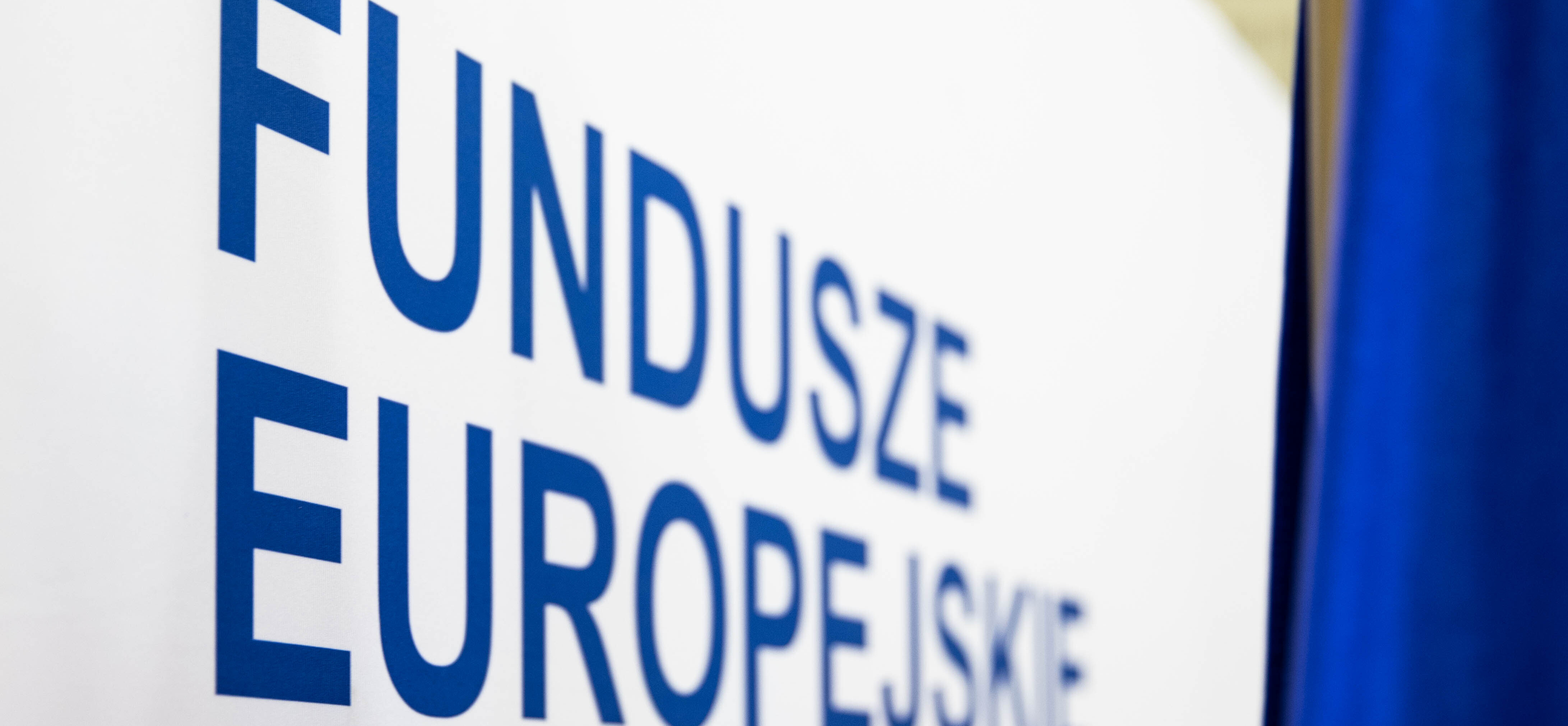 Biała ścianka z niebieskim napisem Fundusze Europejskie