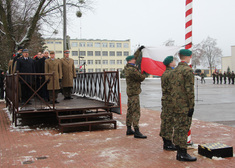 Żołnierze wciągają flagę na maszt, za nimi  stoją zaproszeni goście i mężczyźni w mundurach