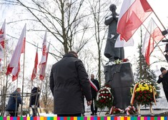 Mężczyzna z pochyloną głową przed pomnikiem ks. Popiełuszki, w tle flagi biało-czerwone