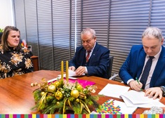 Trzy osoby przy stole. Dwaj mężczyźni podpisują umowę, obok przy stole siedzi kobieta