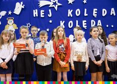 Dzieci stoją w dwóch rzędach przed niebieską ścianką. Jedno z nich trzymna w dłoni mikrofon