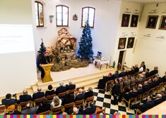 widok z góry salę, szopka bożonarodzeniowa po lewej choinka, po prawej duchowny przy mównicy, goście na krzesełkach
