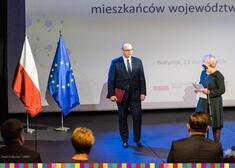 Sekretarz Województwa, po lewej stronie flagi Polski i Unii Europejskiej, po prawej kobieta przy mikrofonie 