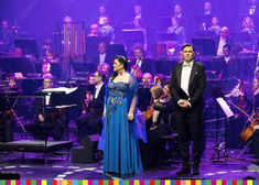 Kobieta w długiej niebieskiej sukni i mężczyzna we fraku, w tle muzycy orkiestry 