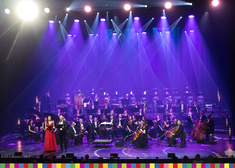 Kobieta w czerwonej długiej sukni i mężczyzna we fraku, za nimi orkiestra opery