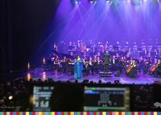 Kobieta w niebieskiej sukni, za nią wszyscy muzycy orkiestry podczas gry