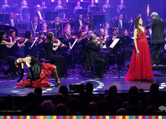 Kobieta w czerwonej sukni w podczas śpiewu, po lewej tancerka w pozie tanecznej, za nimi orkiestra