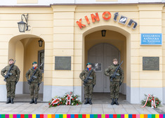 Kwiaty złożone pod tablicą przy kinie Ton. Obok stoją żołnierze.