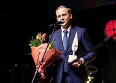 Mężczyzna przy mikrofonie trzyma w rękach kwiaty i statuetkę