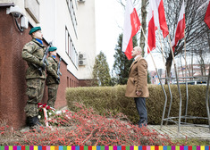 Mężczyzna stoi pod biało-czerwonymi flagami przed tablicą i dwoma żołnierzami