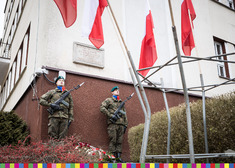 Żołnierze stoją na warcie pod tablicą. Przed nimi wiszą flagi