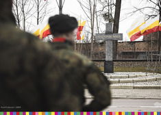 Między flagami stoi pomnik krzyż. Na pierwszym planie widać żołnierzy