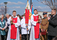 Ministranci trzymają świece i krzyż. Obok stoją ludzie w mundurach