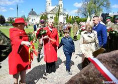 Wiesława Burnos stoi przed pomnikiem, trzyma kwiaty. Wokół uczestnicy wydarzenia. 