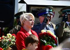 Wiesława Burnos z kwiatami w rękach, za nią stoją salutujący żołnierze. 