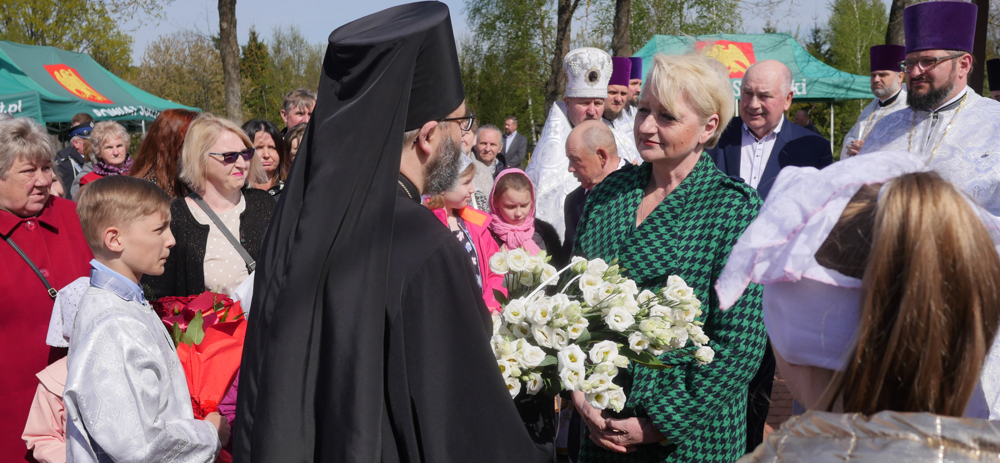 Duchowny trzyma kwiaty przed Wiesławą Burnos, wokół uczestnicy wydarzenia