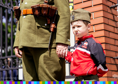 Chłopczyk w furażerce z orłem trzyma dłoń dorosłego w mundurze