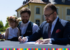 Mężczyzna podpisuje umowę, obok siedzi mężczyzna z dzieckiem