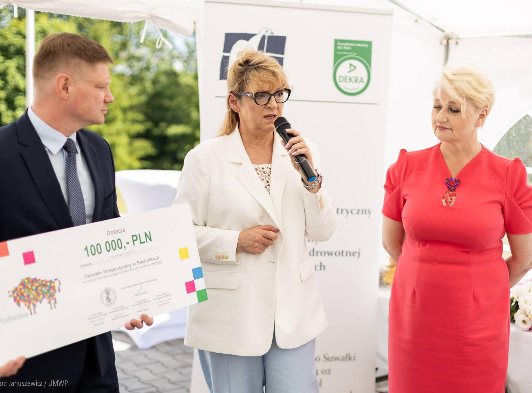 Kobieta stoi między dwoma członkami zarządu i trzyma w dłoni czek na kwotę 100 tys. zł