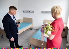 Wiesława Burnos, członek zarządu trzymająca bukiet z kwiatami oraz Marek Malinowski, członek zarządu przebywają w pokoju z łózkami