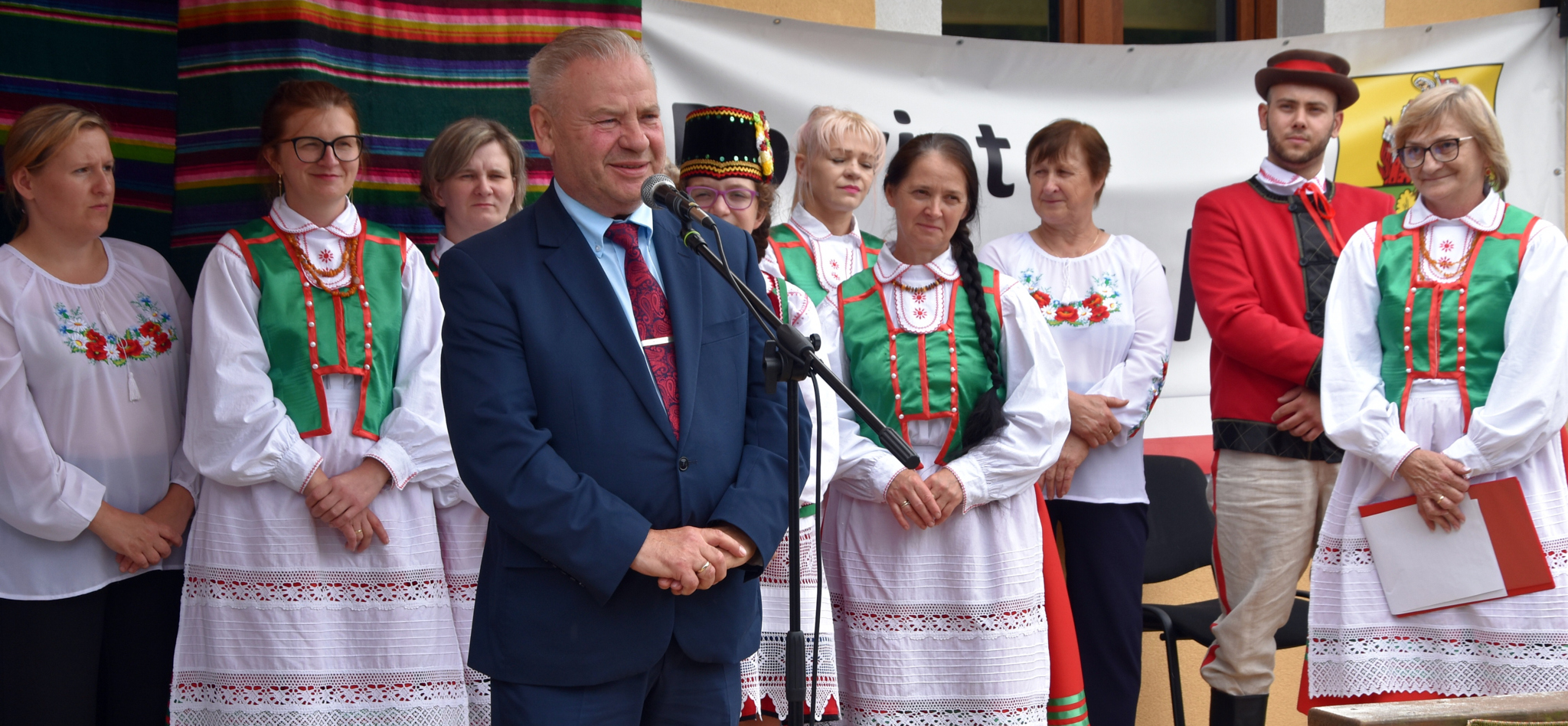 Przemawiający wicemarszałek Marek Olbryś przez mikrofon, a za nim stoją kobiety ubrane w ludowe stroje.
