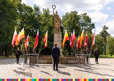 Wicemarszałek Sebastian Łukaszewicz stoi przed pomnikiem przy którym wiszą flagi i stoi asysta honorowa