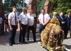 Mężczyźni i kobiety stoją na placu, przed nimi wieńce ze słomy