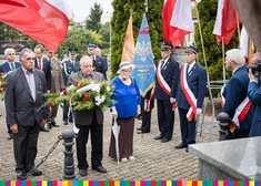 Dwie osoby stojące przed pomnikiem, jedna trzyma kwiaty