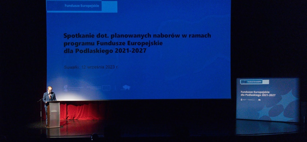 Wielki ekran ze slajdem oraz mężczyzna stojący za mównicą.