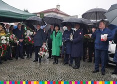 Wiesława Burnos, członek zarządu województwa stoi pod parasolem, po lewej mężczyzna w garniturze, trzyma w ręku kwiaty, po prawej stronie mężczyźni w mundurach, jeden z nich trzyma lampion