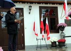 Wypowiedź oficjela na tle tablicy memoratywnej z polskimi flagami w tle