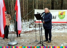 Wicemarszałek Marek Olbryś przed mikrofonem, w tle widać las, po lewej stronie flagę Polski