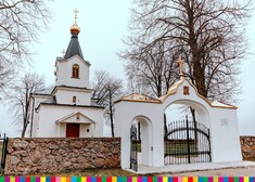 Biała cerkiew z jedną wieżą, kamienny mur odchodzący od bramy z czarną, metalową furtką