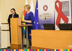 Kobieta stoi na mównicy, obok niej druga kobieta, za nimi flagi: województwa, Polski i Unii Europejskiej 