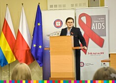 Kobieta stoi na mównicy, po lewej stronie widoczne flagi: województwa, Polski i Unii Europejskiej