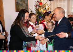Wicemarszałek Marek Olbryś wręcza kobiecie bombkę i pocztówkę świąteczną, w tle widać dzieci w strojach ludowych i choinkę