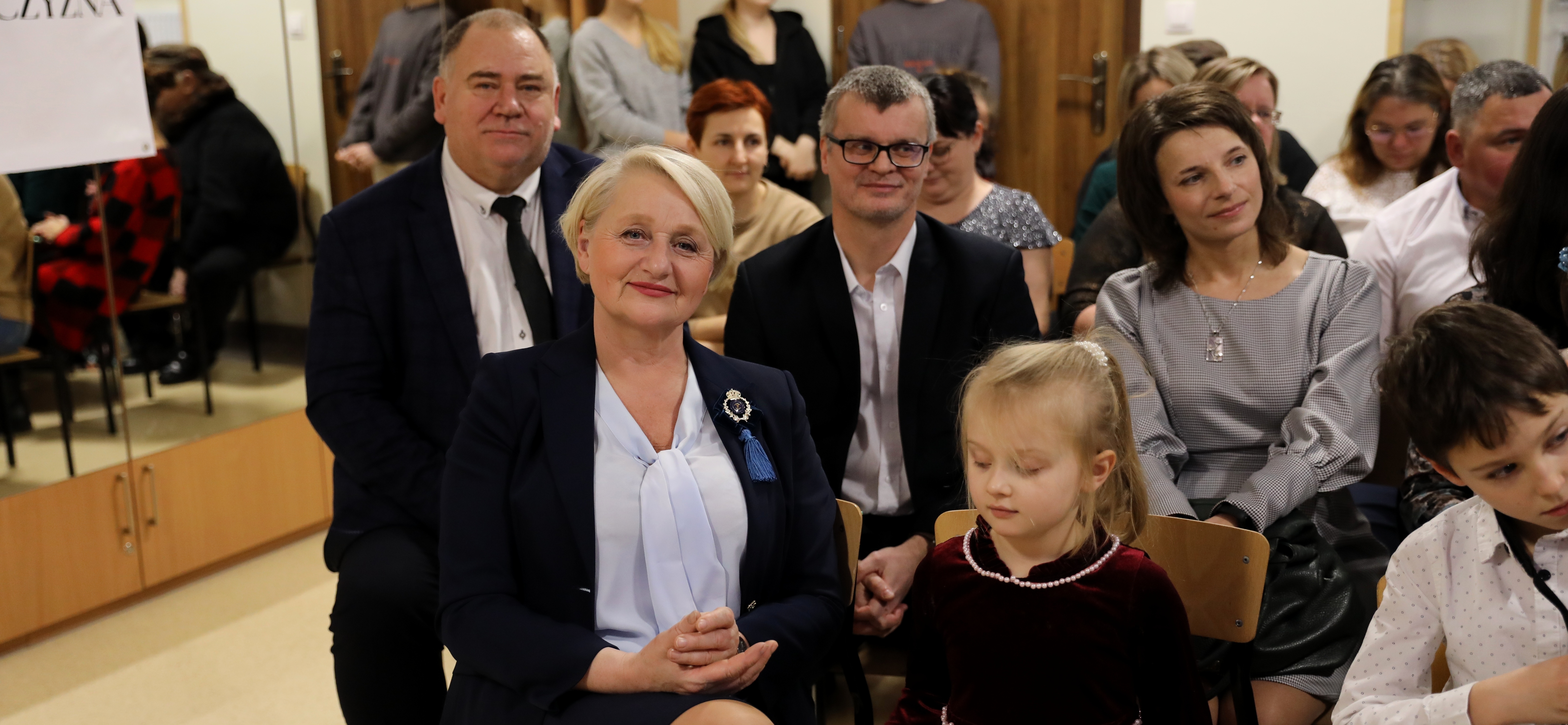 Uśmiechająca się Wiesława Burnos-członek zarządu i weseli widzowie siedzący na krzesłach