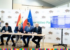 Czterej mężczyźni podpisują na stole dokumenty, w tle znajdują się flagi województwa podlaskiego, Polski i Unii Europejskiej