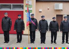 Sześciu mundurowych, jeden z nich trzyma sztandar, stoją przed budynkiem jednostki OSP 