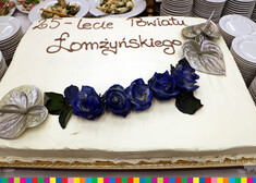 Biały tort z fioletowymi różami i napisem 25- lecie Powiatu Łomżyńskiego